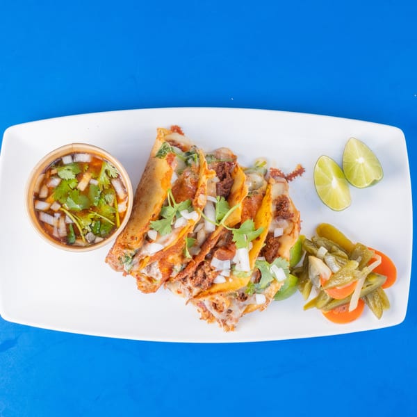 Four cheesy birria tacos with consommé, lime wedges, and pickled vegetables on a white plate.