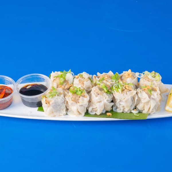 A white plate of steamed dumplings with green onions, fried garlic, lemon, and two dipping sauces.