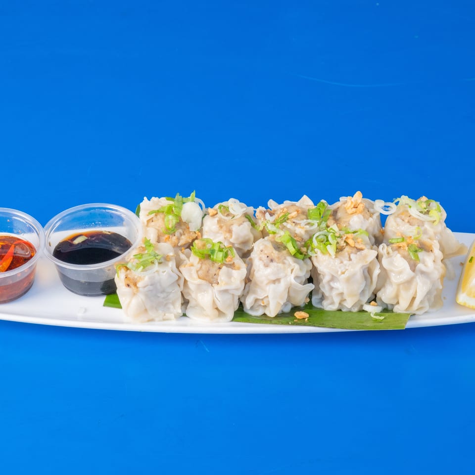 A white plate of steamed dumplings with green onions, fried garlic, lemon, and two dipping sauces.