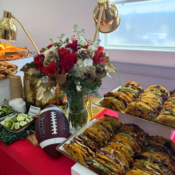 A buffet table with tacos, fried chicken, limes, a floral arrangement, and a football.