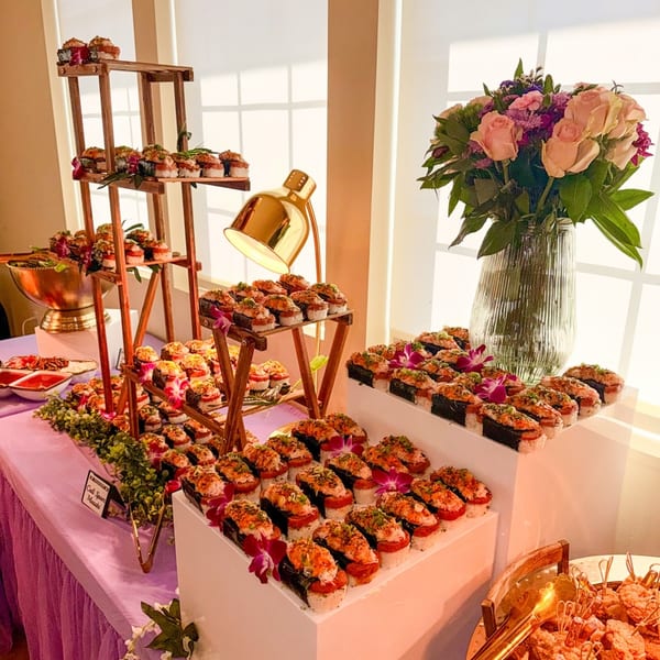 A buffet table with tiered displays of sushi, a golden lamp, and a vase of pink roses.