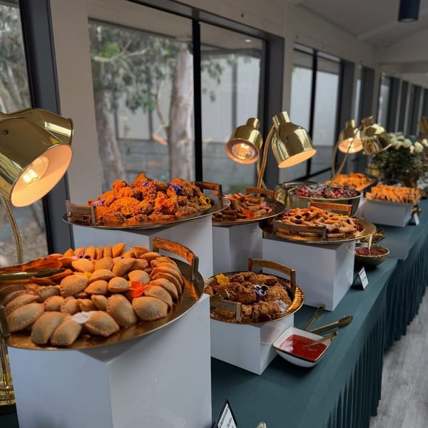 A long buffet table with various appetizers, including fried chicken, empanadas, and spring rolls, under heat lamps.