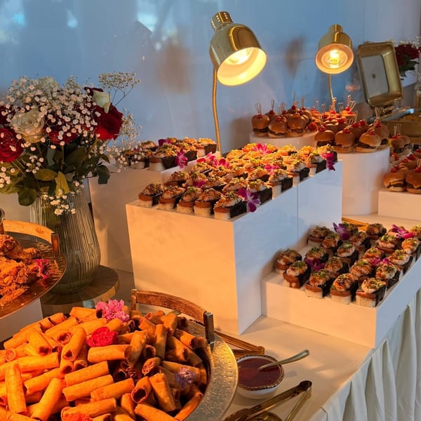 A buffet table laden with spring rolls, sushi, and mini burgers, illuminated by golden lamps.