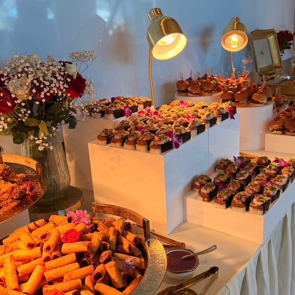 A buffet table laden with spring rolls, sushi, and mini burgers, illuminated by golden lamps.
