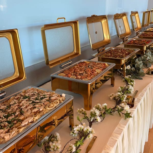 A buffet line with golden chafing dishes filled with pasta, shrimp, and other dishes, adorned with floral garlands.