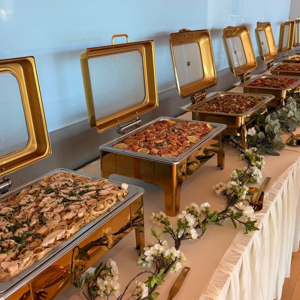 A buffet line with golden chafing dishes filled with pasta, shrimp, and other dishes, adorned with floral garlands.