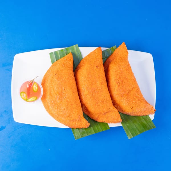 Three golden-fried empanadas on banana leaves with chili sauce, on a white plate against a blue background.